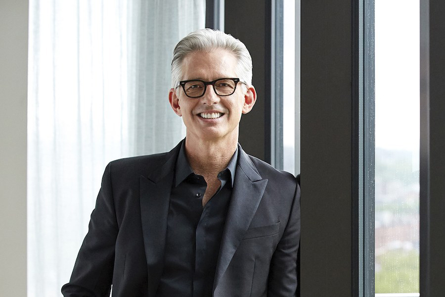 Smiling man with gray hair wearing glasses and a black suit standing by a window at an event venue space.