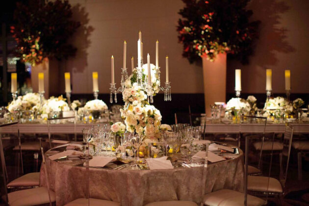 Elegant banquet table setting with floral centerpieces and candlesticks in a dimly lit event venue space.