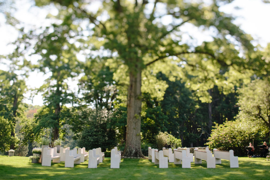 A serene event venue space with white chairs arranged for an occasion under the shade of a large tree.