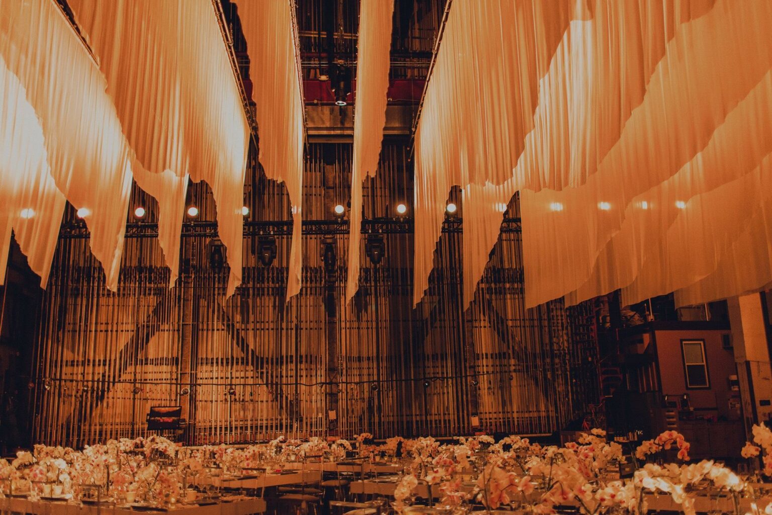 Elegant banquet setup arranged by event planners, with tables adorned with flowers and warm lighting under a canopy of flowing drapery.