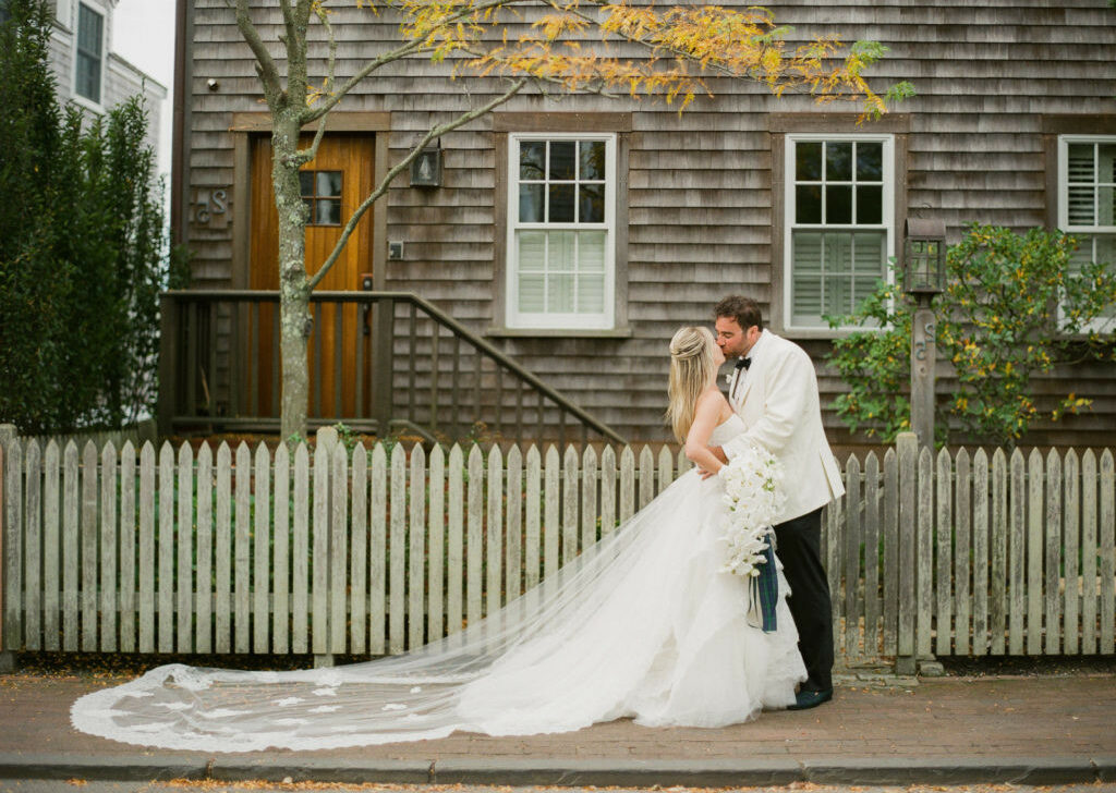 Bride and groom sharing a kiss on a sidewalk with a rustic wooden house and a white picket fence in the background, perfectly orchestrated by an event planning company.