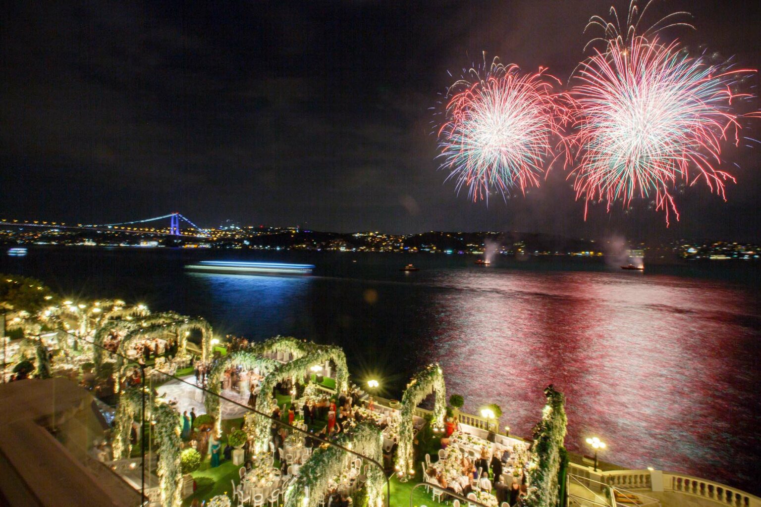 fireworks exploding in the air over water next to an outdoor event reception.