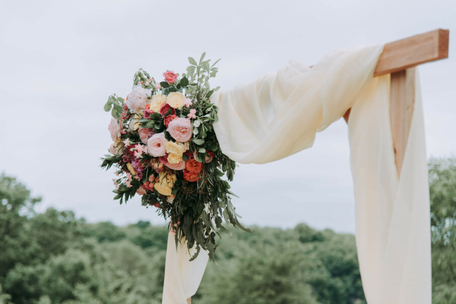 A lush floral arrangement attached to a wooden arch draped with a light fabric outdoors, designed by an event planning company.