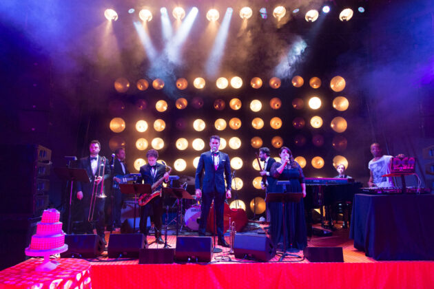 Live band performing on stage with colorful stage lighting and a festive atmosphere at an event venue space.