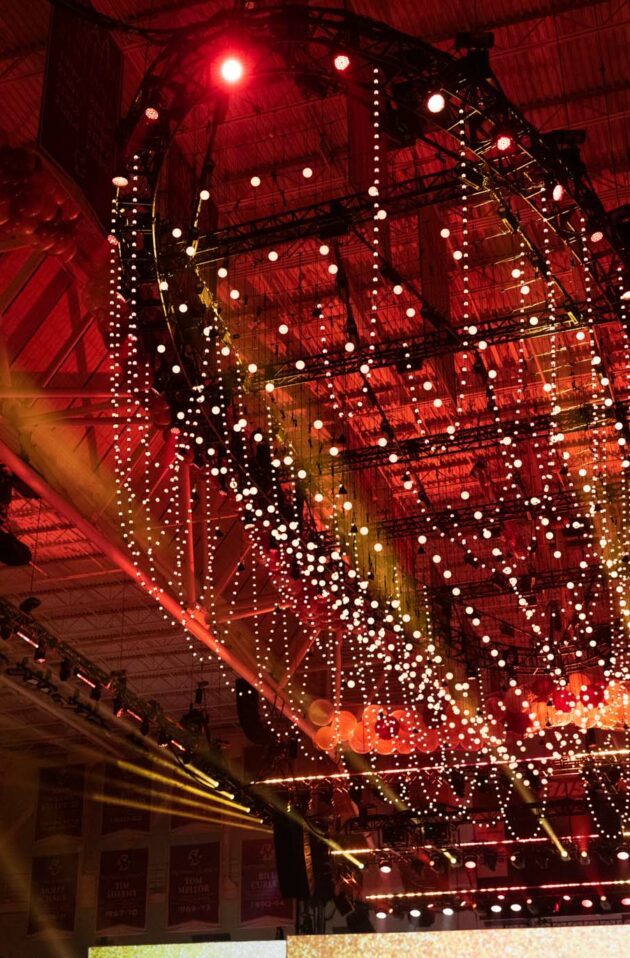 Event planners arranged string lights hanging from a large spherical frame inside a dimly lit indoor arena with spectators.