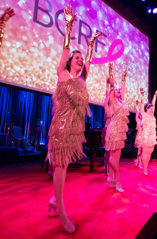 Performers in sparkling costumes dancing on stage at an event with the word "broad" projected in the background, organized by a renowned event planning company.