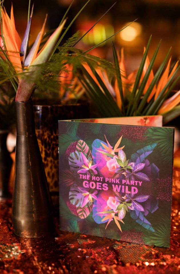 A vibrant party invitation, crafted by a leading event planning company, with tropical design elements, positioned next to a floral arrangement on a sequined surface.