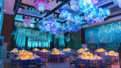 An event venue space decorated with blue and purple balloons, illuminated tables, and hanging chandeliers.