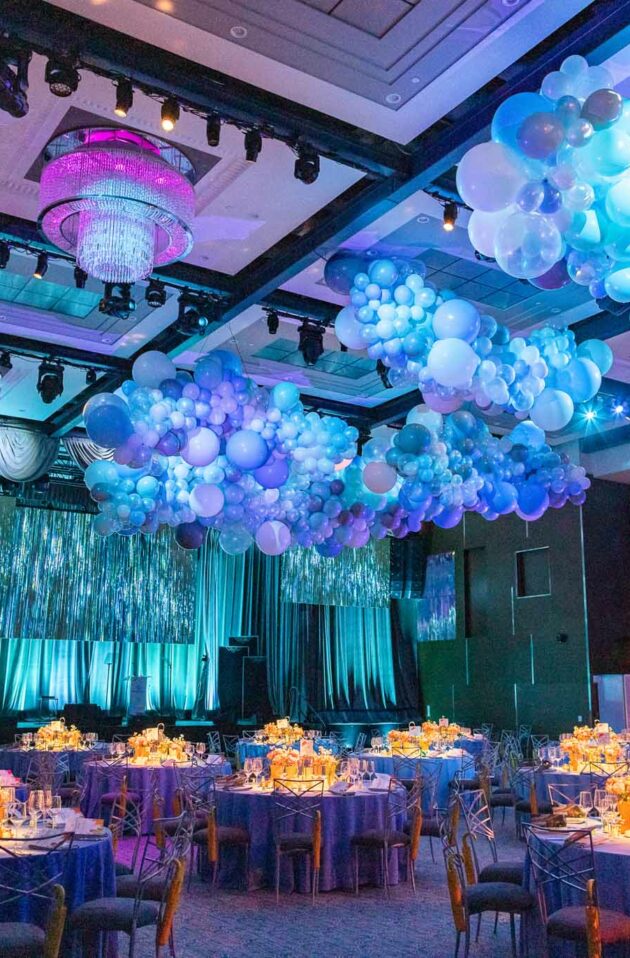 An event venue space decorated with blue and purple balloons, illuminated tables, and hanging chandeliers.