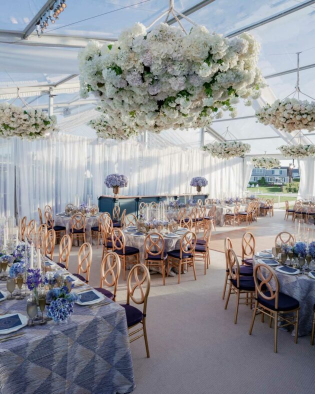 An elegant outdoor event venue space with floral decorations hanging from the ceiling, arranged tables with blue accents, and transparent tent walls.