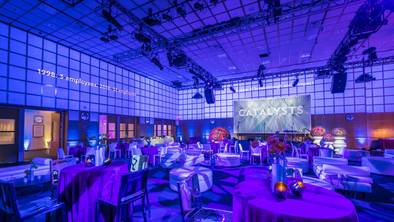 An empty event venue space decorated with blue lighting and set tables, featuring a stage with a prominent "catalysts" backdrop.