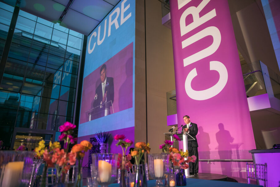 A man speaking at a podium during an event planned by an event planning company, with the word "cure" projected on a screen in the background.