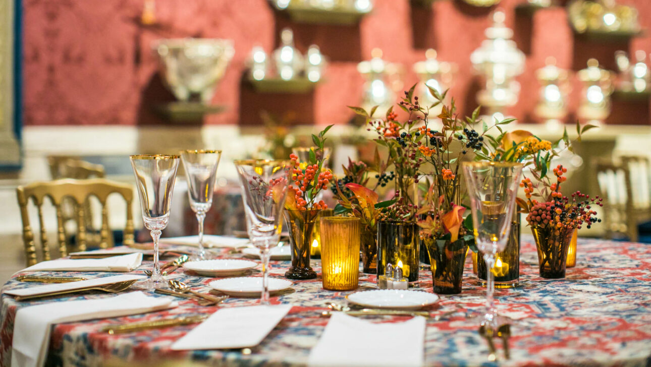 Elegant dining table set with floral centerpieces, fine glassware, and ornate silver in a richly decorated event venue space.