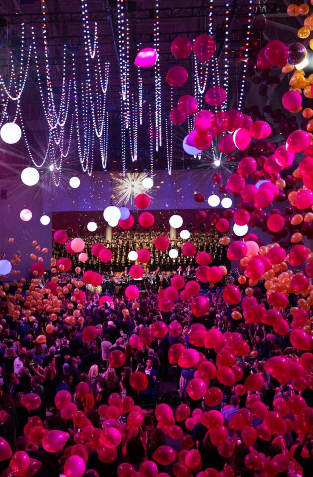 A vibrant event planned by event planners, with a crowd and a burst of red balloons under a canopy of lights.