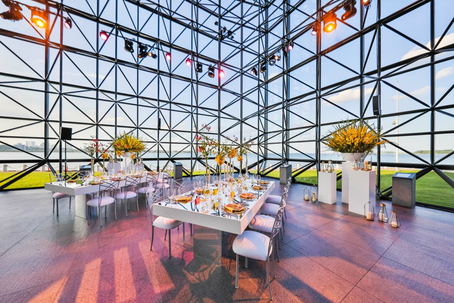 A modern event venue space with a geometric glass facade, elegantly set dining tables, and a view of the water and skyline.