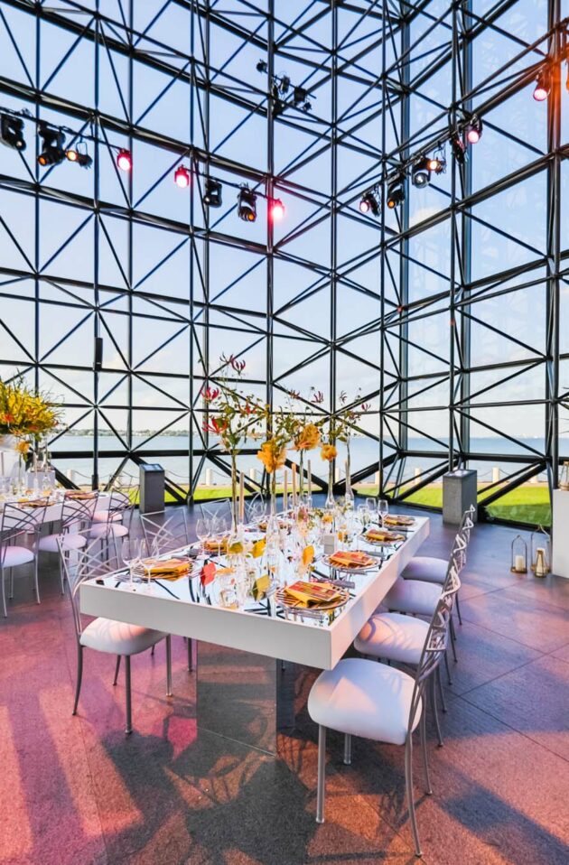 A modern event venue space with a geometric glass facade, elegantly set dining tables, and a view of the water and skyline.