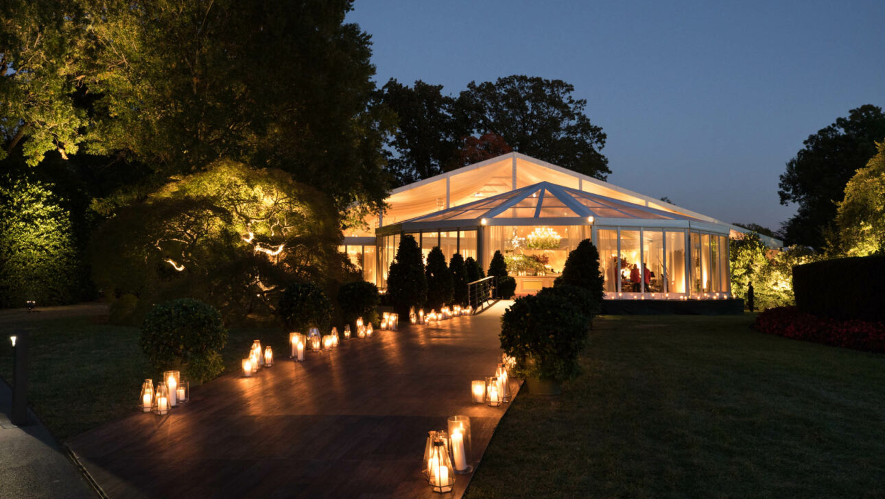 An illuminated, transparent marquee set up for an evening event in a garden adorned with pathway lights by an expert event planning company.