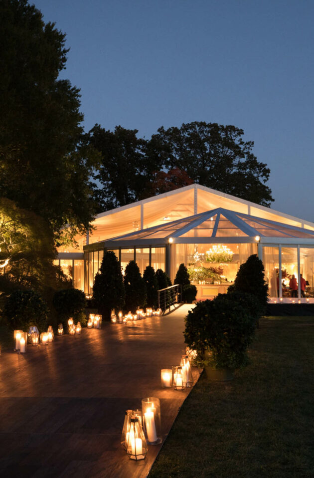 An illuminated, transparent marquee set up for an evening event in a garden adorned with pathway lights by an expert event planning company.