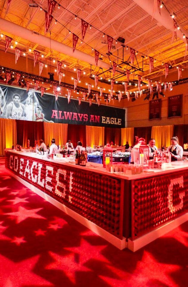 A decorated event venue space with red lighting, a stage set up for a gathering, and tables with tablecloths, featuring a large "cornell" sign in the foreground.