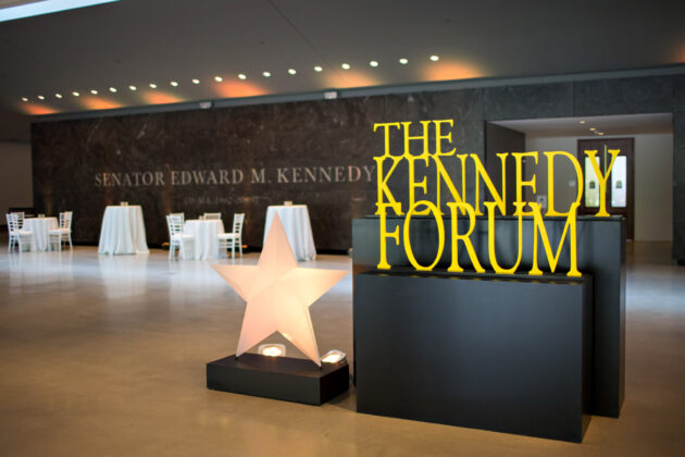 An illuminated sign for "the kennedy forum" with a large star symbol, set up in an event venue space with chairs and tables.