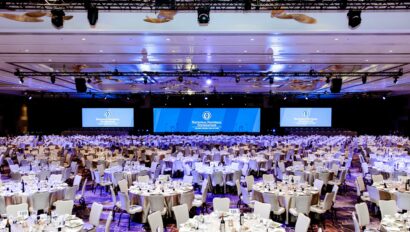 A large event venue space set up for a formal event with multiple dining tables and a stage with screens displaying a logo.