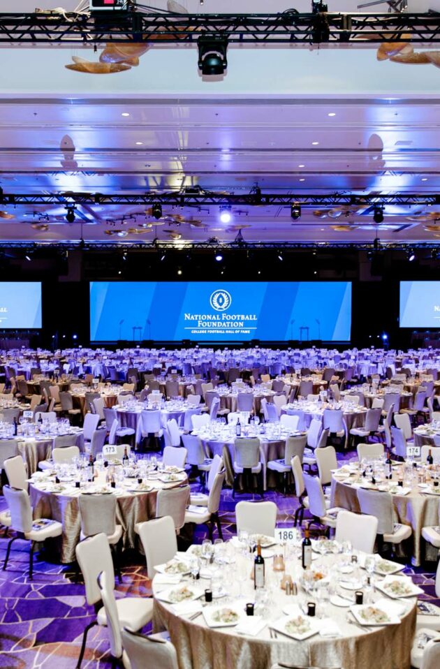 A large event venue space set up for a formal event with multiple dining tables and a stage with screens displaying a logo.