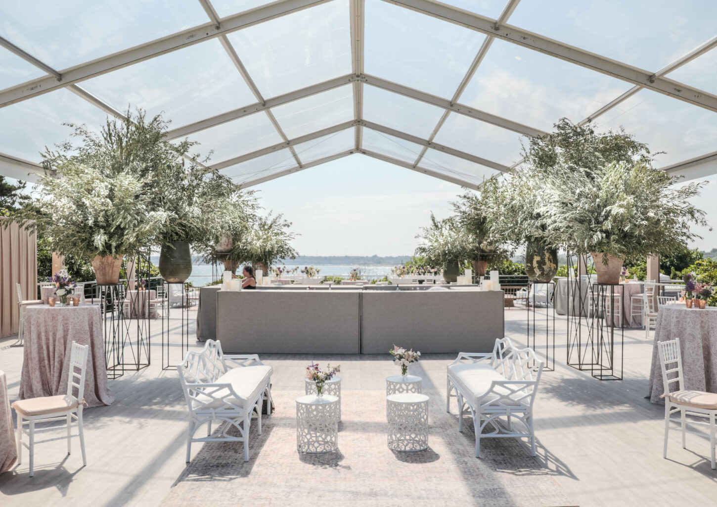 An elegant outdoor event setup under a clear marquee, with tables, chairs, and potted plants, curated by a premier event planning company, facing a waterfront view.