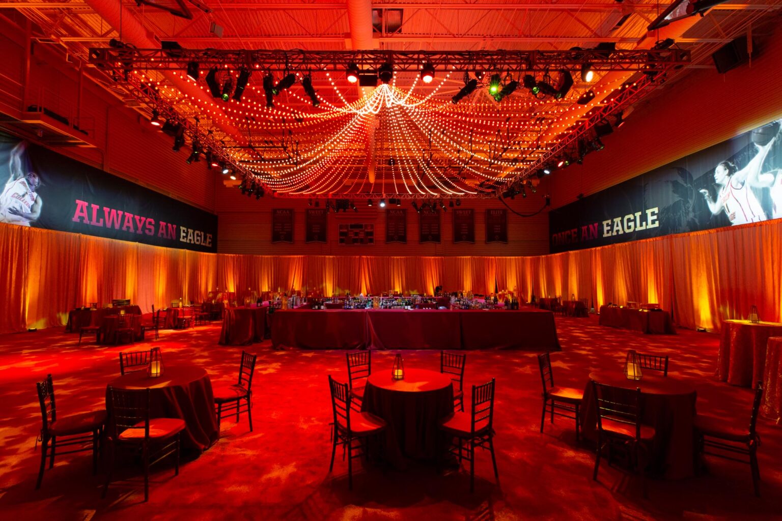 A banquet hall prepared by an event planning company for an event with tables, chairs, and dramatic red lighting with a banner that reads "always an eagle.