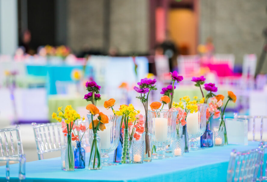 Brightly colored flowers arranged in vases on a table with blue tablecloth, accompanied by lit candles, set for an event by an event planning company.