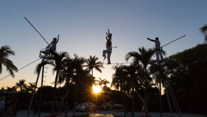 Silhouetted tightrope walkers perform at sunset with tropical trees in the background, an unforgettable spectacle organized by skilled event planners.