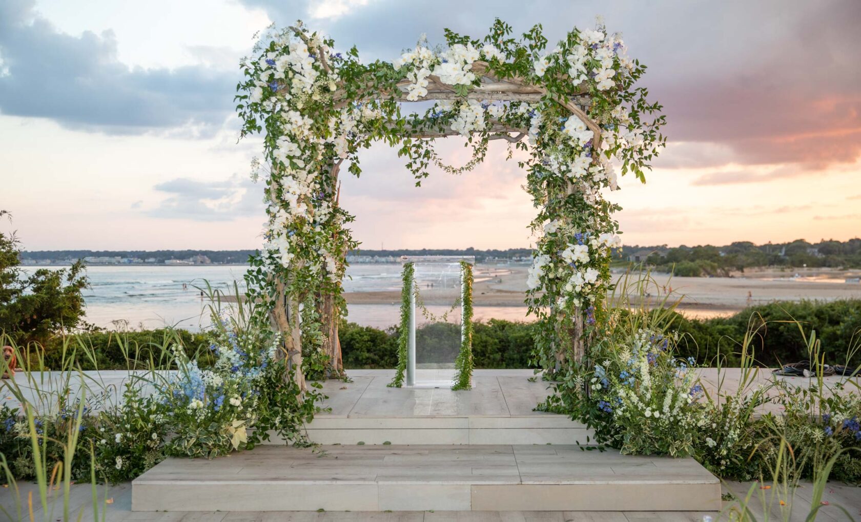 A floral-decorated wedding arch, organized by an event planning company, overlooking a beach at sunset.