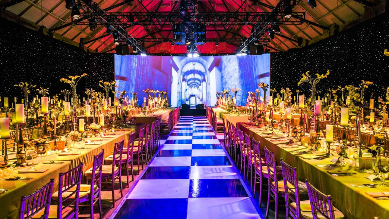 Grand banquet hall set for a formal event by an event planning company, with a blue and white checkerboard aisle, decorated tables, and a large projected image on the wall.