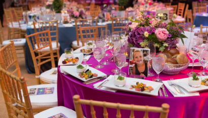 An elegantly set banquet table at an event venue space, with a vibrant pink tablecloth, floral centerpieces, and place settings that include photographs.