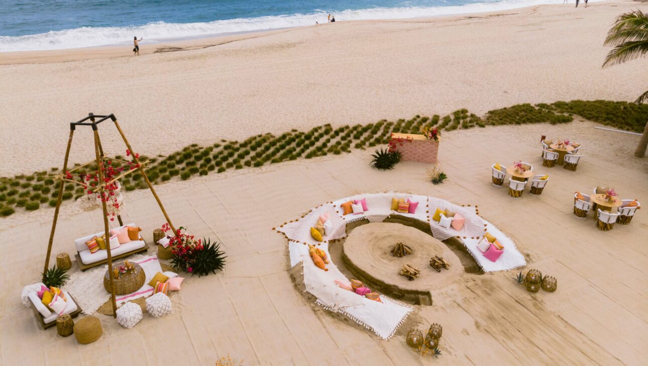 Aerial view of a beach wedding setup organized by an event planning company, with circular seating arrangement and a floral arch.