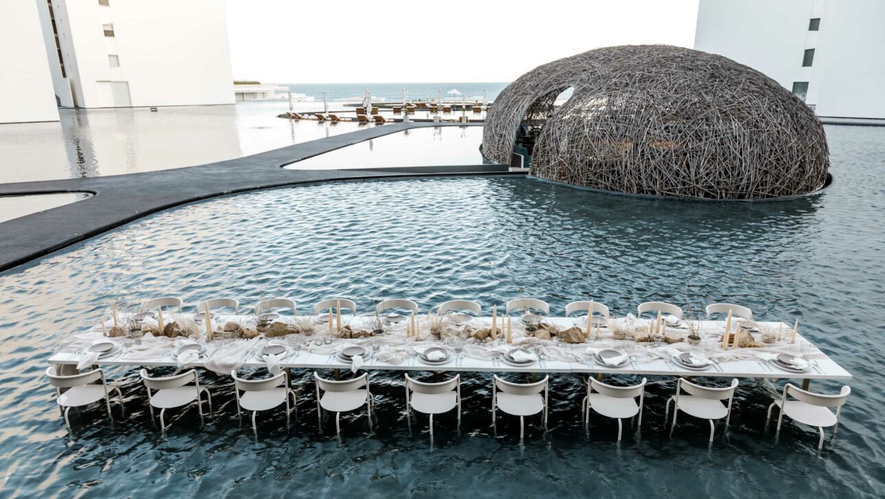 An outdoor dining table, set for a meal by an event planning company, partially submerged in a pool, with a woven dome structure in the background.