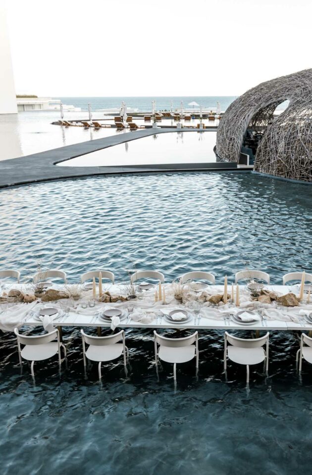 An outdoor dining table, set for a meal by an event planning company, partially submerged in a pool, with a woven dome structure in the background.