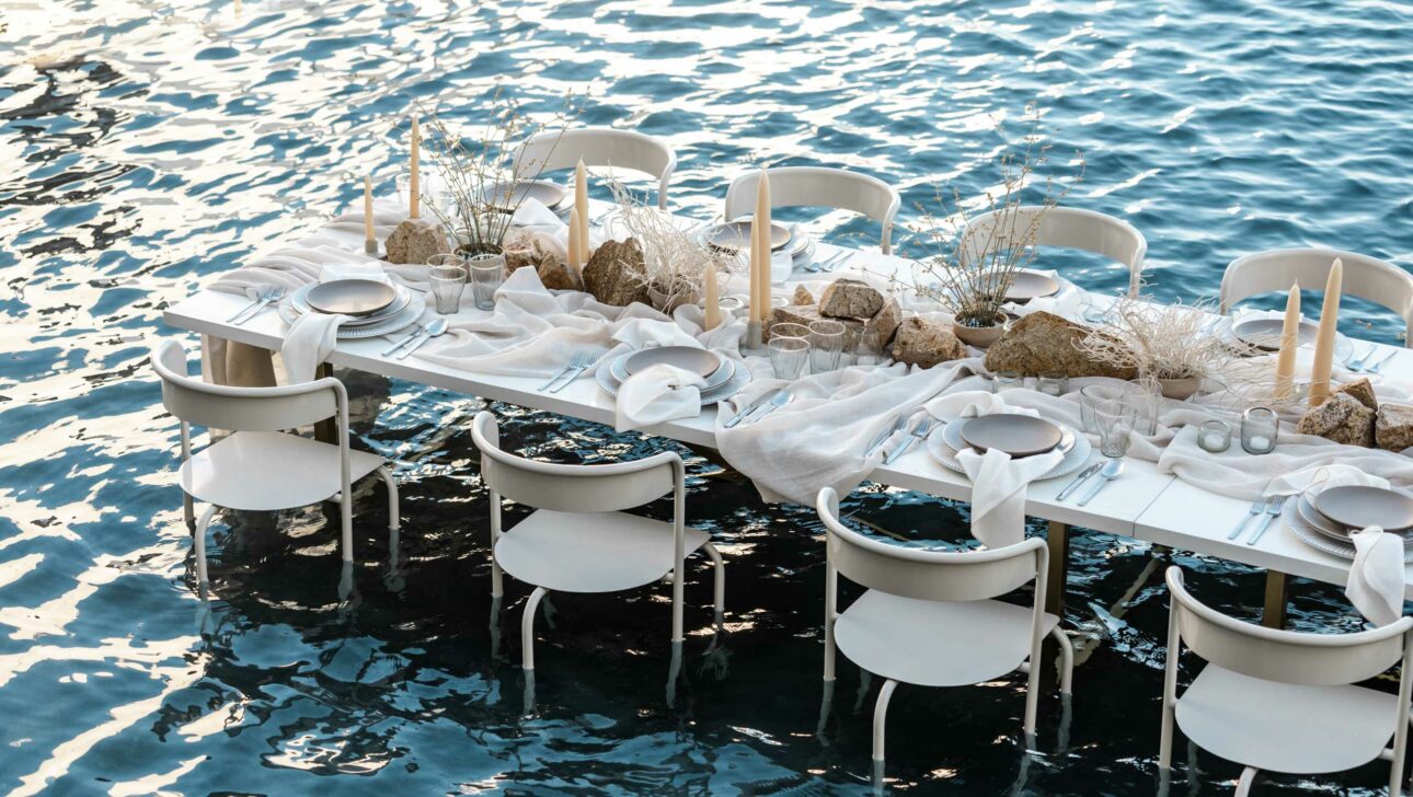 A dining table set with white decor, arranged by an expert event planning company, floating on a calm blue body of water at dusk.