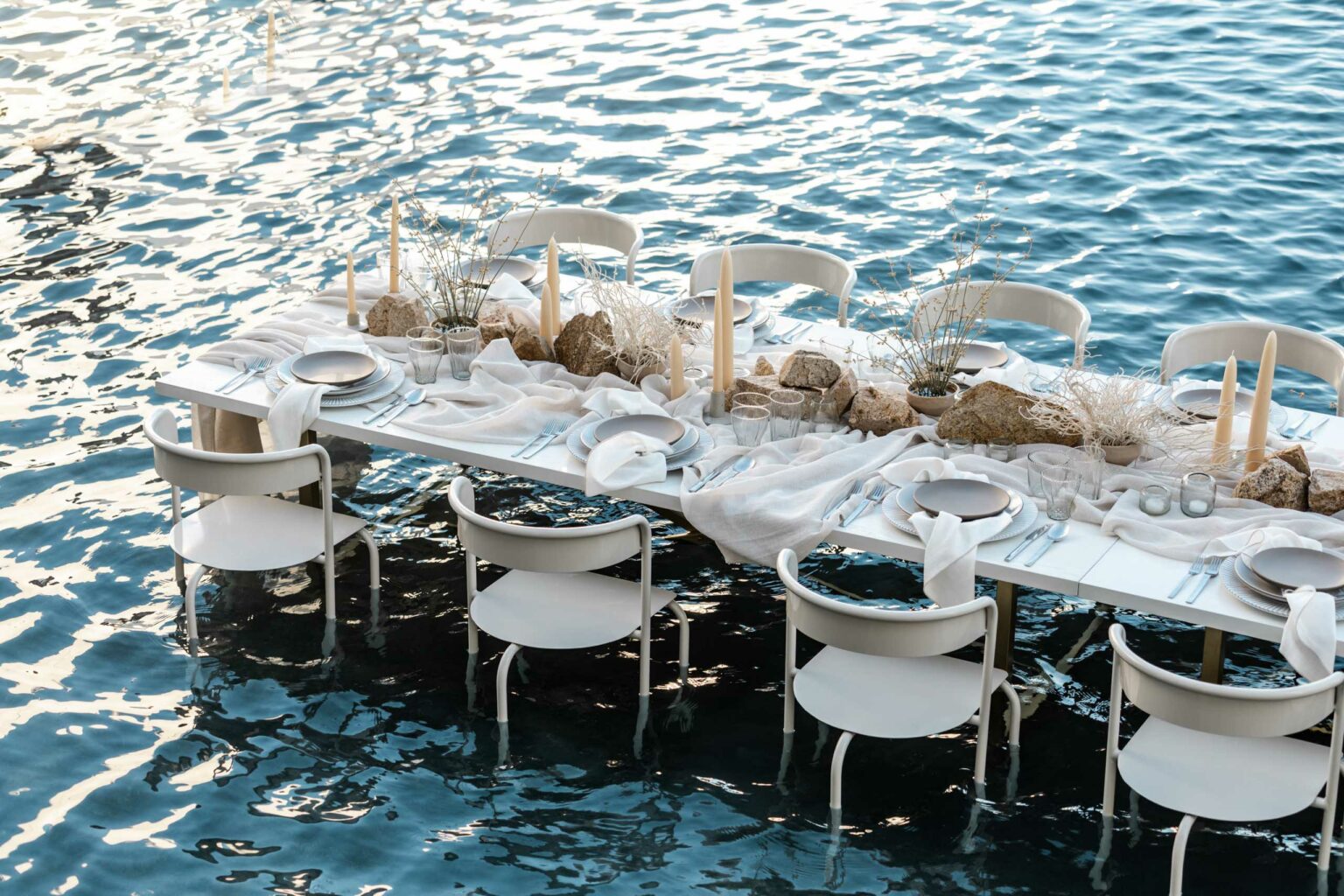 A dining table set with white decor, arranged by an expert event planning company, floating on a calm blue body of water at dusk.