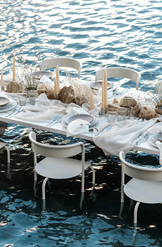 A dining table set with white decor, arranged by an expert event planning company, floating on a calm blue body of water at dusk.