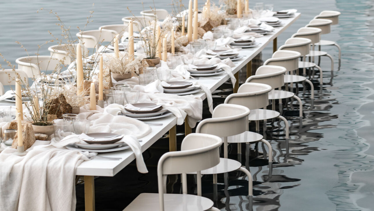 Elegantly set dining table, orchestrated by skilled event planners, partially submerged in water with chairs and decorative grass centerpieces.