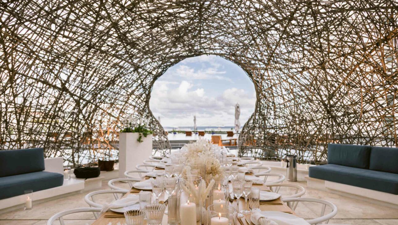 Elegant outdoor dining setup with a long table, decorative flowers and candles, designed by a leading event planning company, under an intricate woven canopy with a view of the sky.