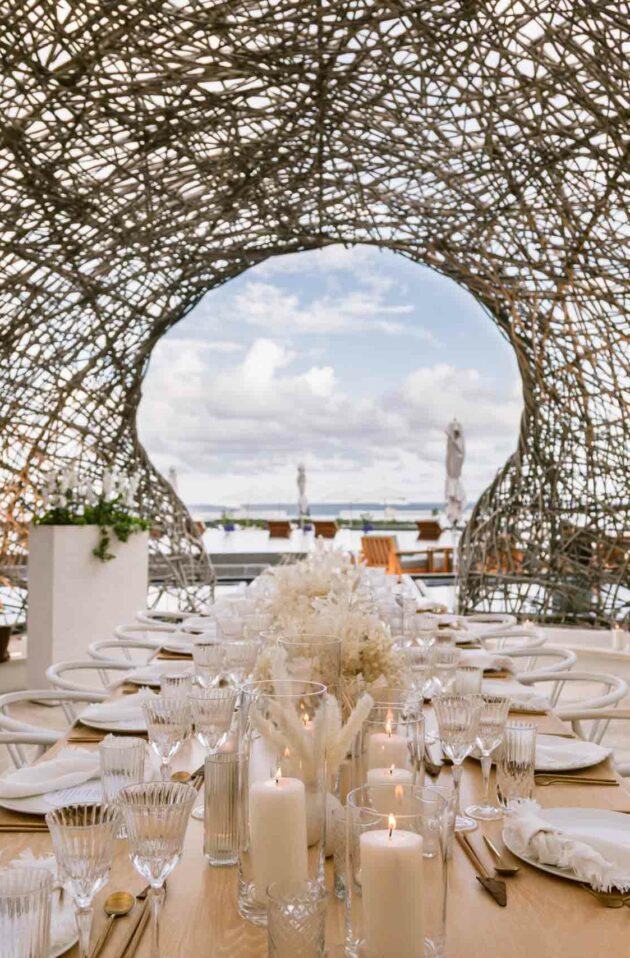 Elegant outdoor dining setup with a long table, decorative flowers and candles, designed by a leading event planning company, under an intricate woven canopy with a view of the sky.