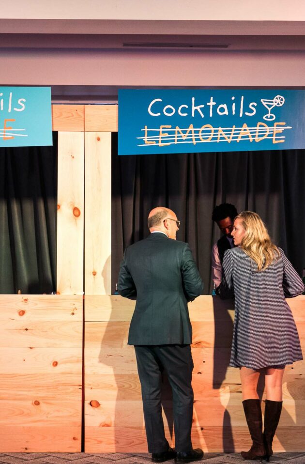 Three event planners at a makeshift wooden counter with signs overhead reading "cocktails lemonade.