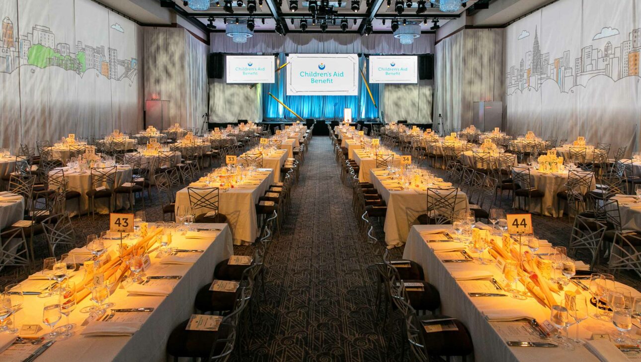 Banquet hall set up for a formal event by an event planning company, with tables, chairs, and a stage with a "children's aid benefit" banner.