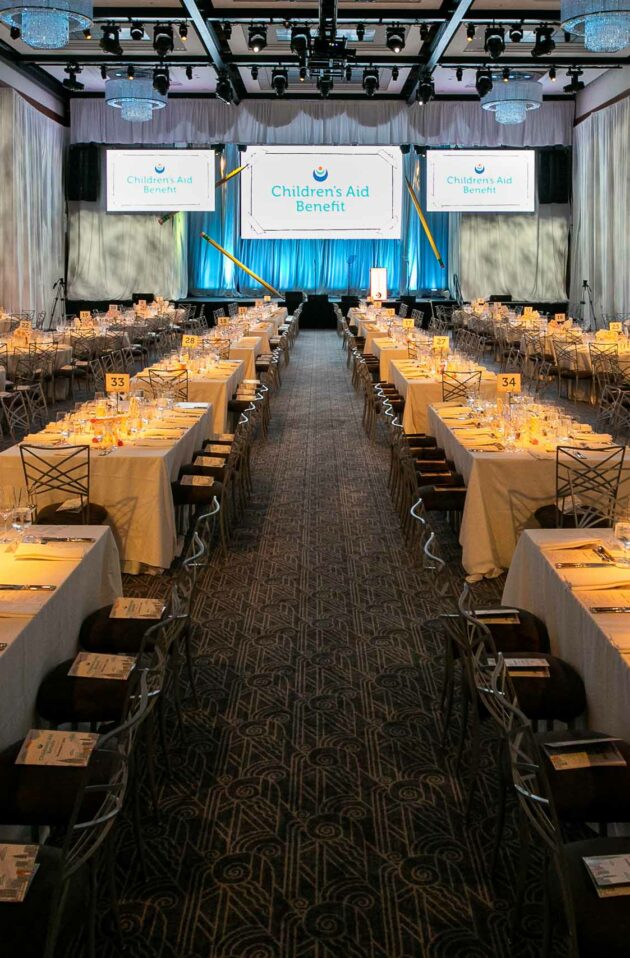 Banquet hall set up for a formal event by an event planning company, with tables, chairs, and a stage with a "children's aid benefit" banner.