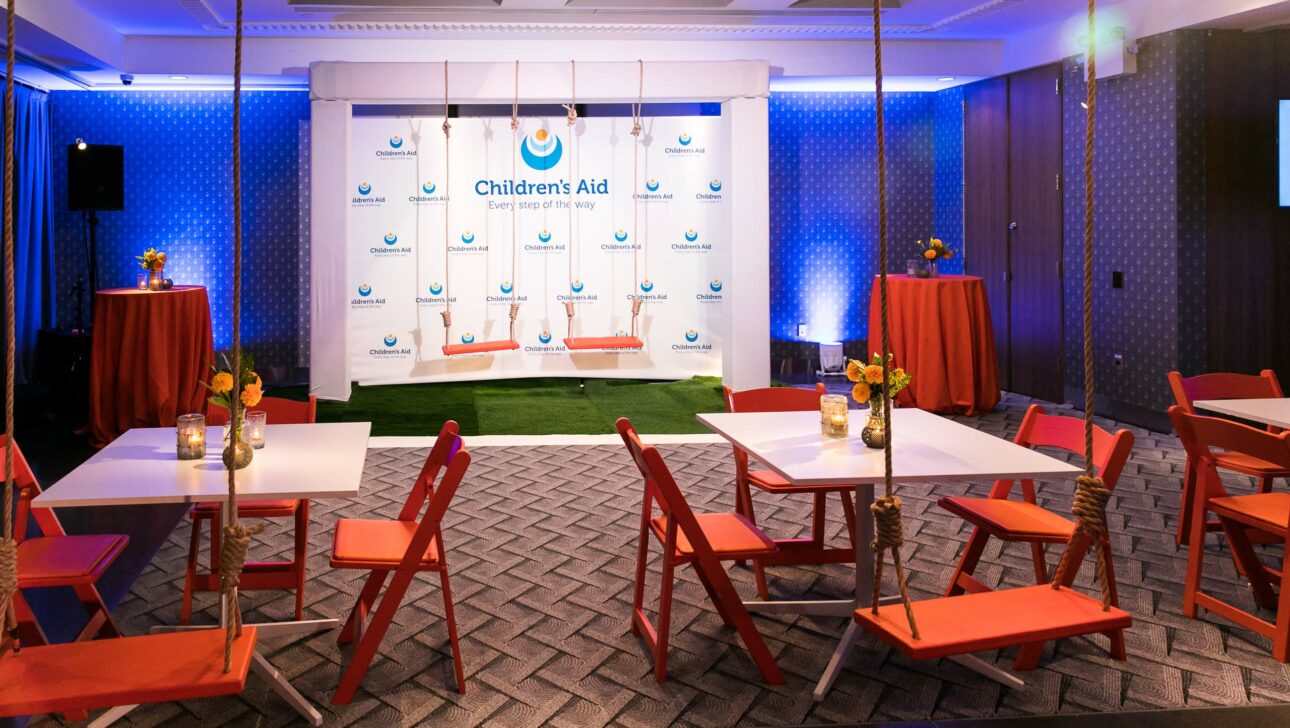 Event venue space set up for a gathering with branded backdrop, cocktail tables, and red chairs.