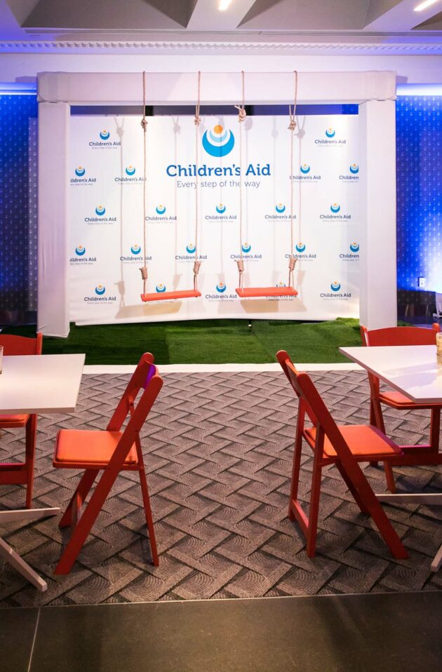 Event venue space set up for a gathering with branded backdrop, cocktail tables, and red chairs.