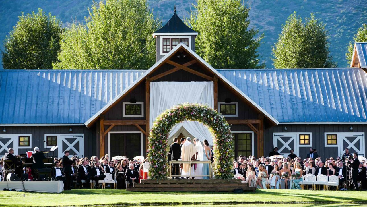 A wedding ceremony organized by event planners is taking place outdoors by a pond, with guests seated and a floral arch at the venue's entrance.