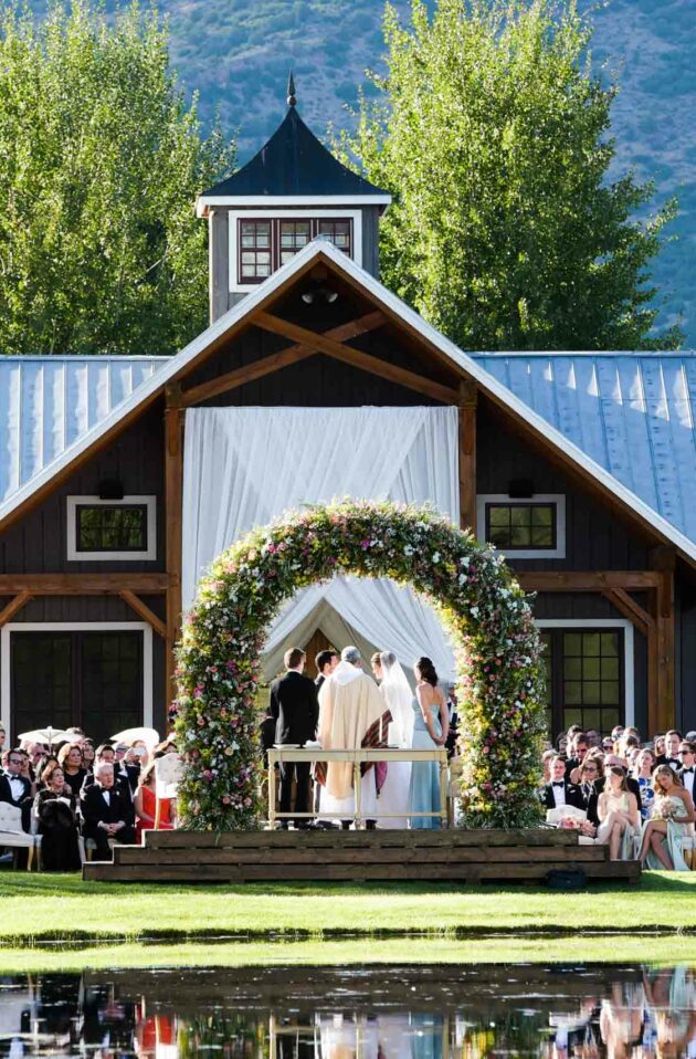 A wedding ceremony organized by event planners is taking place outdoors by a pond, with guests seated and a floral arch at the venue's entrance.