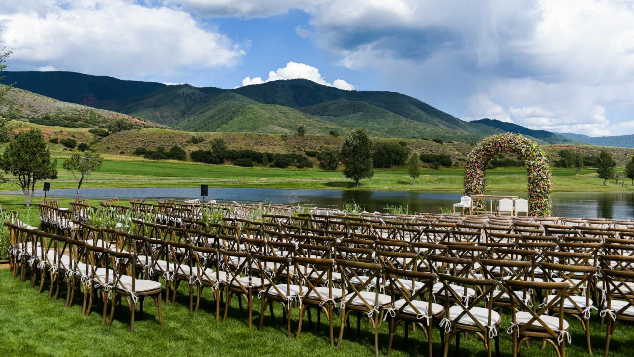 chairs set out for a wedding ceremony outside next to a lake with a floral arch.